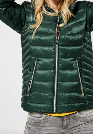 Kvinde iført en mørkegrøn quiltet jakke med sølvfarvede lynlåslommer og lynlås foran, kombineret med lysegrå jeans og en sennepsgul skjorte under.