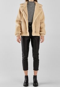 Kobieta ubrana w beżową kurtkę z owczej wełny, czarne skórzane spodnie kończące się nad kostką, czarne botki oraz koszulę w czarno-białe paski, stojąca na szarym tle.