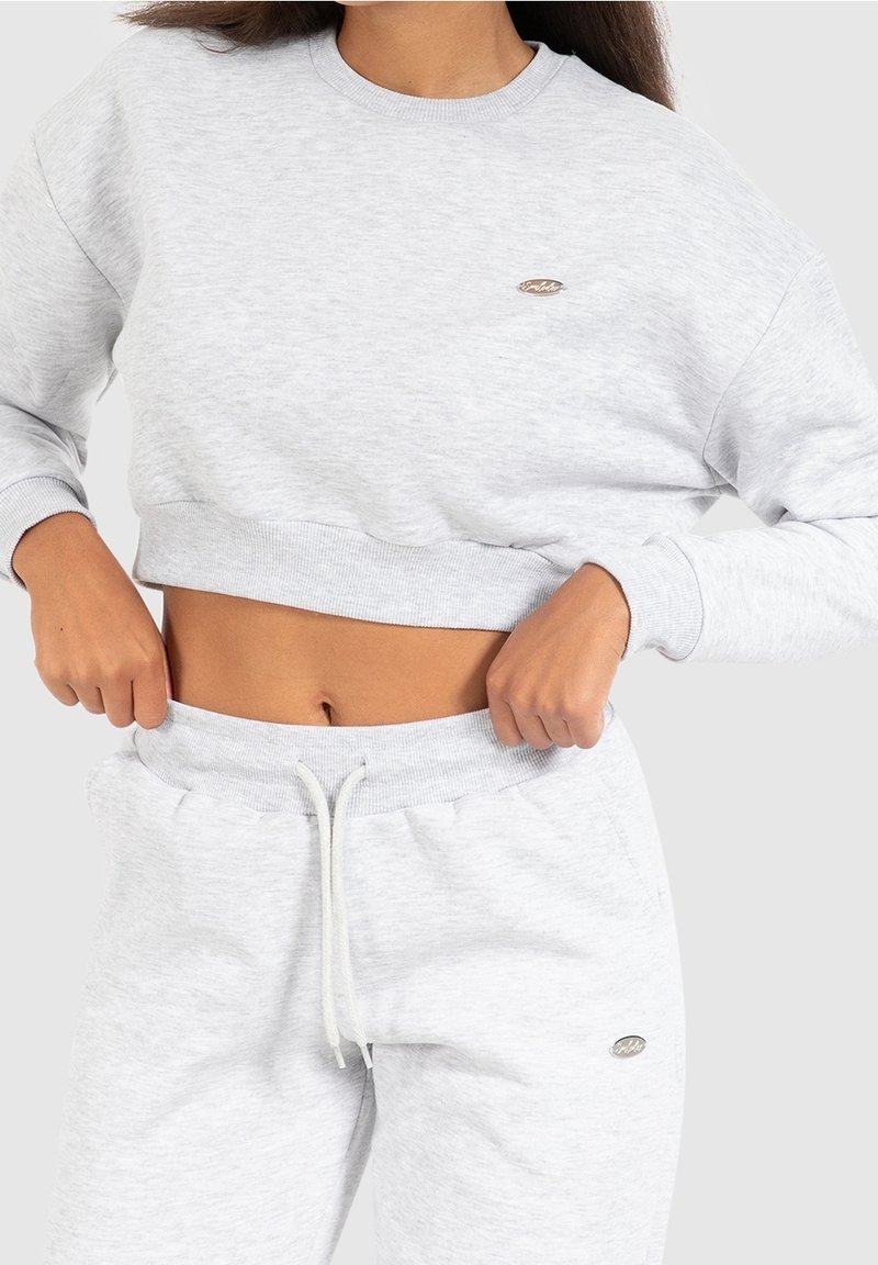 Femme ajustant un sweat-shirt court gris clair et un pantalon de jogging assorti avec un cordon, montrant le ventre nu sur un fond uni.