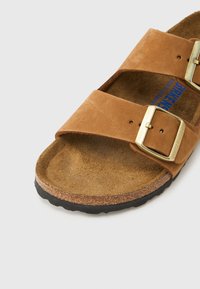 Καφέ σουέντ σανδάλι με δύο φαρδιές λουριά και αγκράφες σε χρυσή απόχρωση. Υφή στο πέλμα, με μεσαία σόλα από φελλό και μαύρη λαστιχένια εξωτερική σόλα.