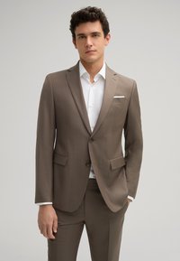 Brauner maßgeschneiderter Anzug mit einer Zwei-Knopf-Verschluss, Reverskragen und zwei Taschen. Begleitet von einem weißen Hemd mit glatter Textur.