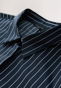 Chemise bleu marine à col avec fines rayures blanches verticales, soigneusement pliée, montrant le col et une partie du rabat de boutons.