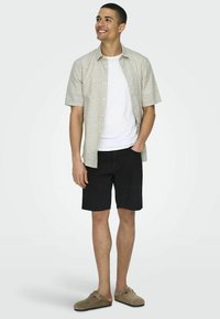Camisa de lino beige claro de manga corta sobre una camiseta blanca, combinada con pantalones cortos negros y zapatos de lona grises, de pie frente a un fondo gris.