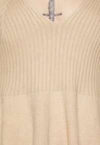 Beżowy sweter z dzianiny, z pionowym prążkowaniem, z dekoltem w szpic i gładką teksturą, z widoczną zmianą koloru przy dolnym końcu.