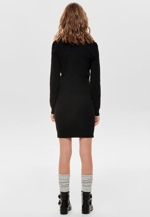 Kobieta z falującymi włosami, ubrana w czarną sukienkę z długim rękawem, szare luźne skarpety i czarne botki, stojąca tyłem na białym tle.