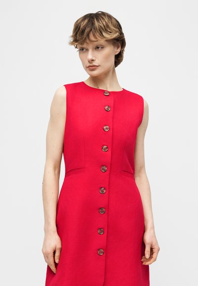 Γυναίκα με κοντά μαλλιά που φοράει κόκκινο φόρεμα χωρίς μανίκια με μια σειρά καφέ κουμπιών μπροστά, σε απλό ανοιχτό φόντο.