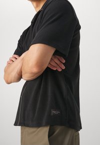 Musta lyhythihainen polo-paita pehmeästä kankaasta rentoa istuvuutta varten, jossa on sivuvetoketju ja pieni etiketti helmassa.