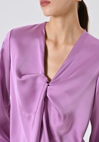 Donna che indossa una blusa lavanda lucida con un dettaglio a nodo attorcigliato sul davanti e una scollatura a V profonda, visibile dal collo fino a metà torace.