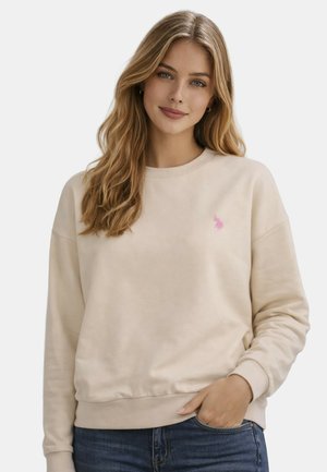 Junge Frau mit langen blonden Haaren, trägt ein beiges Sweatshirt mit einem kleinen pinkfarbenen Logo und blaue Jeans, Hand in der Tasche, lächelt sanft.
