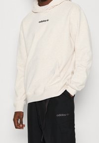 Hellbeige Kapuzenpullover mit einer Fronttasche, ausgestattet mit einem schwarzen "adidas"-Logo auf der Brust und gerippten Bündchen, kombiniert mit dunklen Hosen.