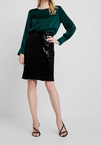 Blusa de satén verde con mangas largas; falda lápiz negra con estampado de leopardo en textura de lentejuelas; tacones altos negros con tiras.