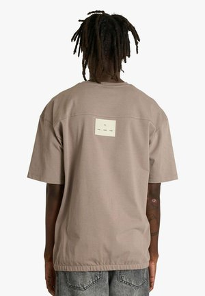 Maglietta a maniche corte in cotone marrone chiaro con collo rotondo e un'etichetta rettangolare sul retro con testo stampato in nero.