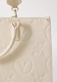 Sac à main en cuir texturé couleur crème, mettant en valeur des motifs floraux embossés et un logo circulaire avec un "V", agrémenté de quincaillerie dorée.