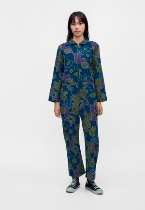 Femme aux longs cheveux foncés, debout, portant une veste à fermeture éclair et un pantalon assortis à imprimé floral bleu et vert avec des baskets montantes noires.