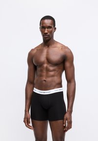 Svarta boxerkalsonger med vit midjeband som har "TOMMY HILFIGER"-logotypen. Slät tyg, åtsittande design, mitt-lår längd.