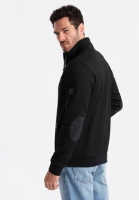 Giacca nera con zip e colletto alto, texture a coste e toppe contrastanti sui gomiti. Presenta tasche laterali e polsini aderenti.