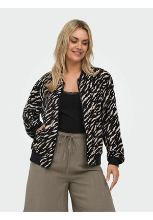 Smilende kvinde iført en sort og beige mønstret lynlåstrøje, sort top og olivengrønne bukser med vide ben og snøre i taljen, poserer mod en hvid baggrund.