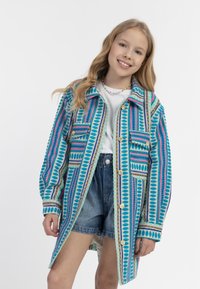 Värikäs ylisuurennettu takki, jossa on sinisiä, vaaleanpunaisia ja turkooseja geometrisia kuvioita, kaksi etupussia ja napitettava etuosa.