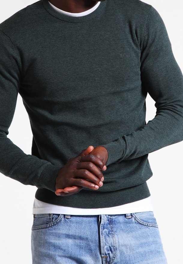 Mężczyzna w ciemnozielonym swetrze z długim rękawem, założonym na białą koszulę, w jasnoniebieskich dżinsach, z rękami złożonymi na brzuchu na jednolitym tle.