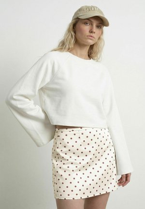 Ung kvinde iført beige kasket, hvid langærmet cropped top og hvid nederdel med brune polkaprikker, poserer med hånden på hoften.