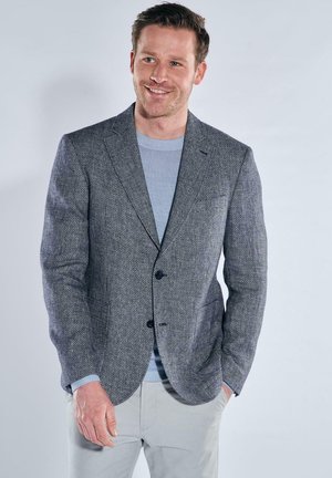 Smiling man wearing a light blue sweater under a textured gray blazer with light-colored pants, standing against a plain light background.