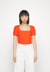 Top curto de manga curta em laranja brilhante, com decote quadrado, combinado com calças de ganga brancas de cintura alta. Textura suave e design justo.