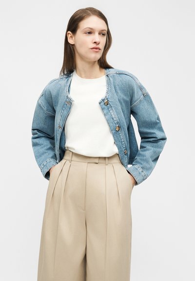 Νεαρή γυναίκα που φοράει ανοιχτό μπλε τζιν μπουφάν με κουμπιά, λευκό πουκάμισο και μπεζ πλισέ παντελόνι ψηλόμεσο, μπροστά σε λευκό φόντο.