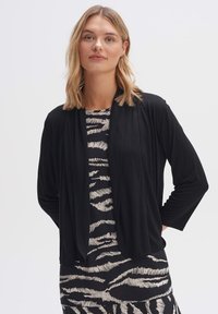 Zwarte cardigan met een gedrapeerde voorkant en driekwartmouwen, gedragen over een gestreepte zwarte en beige jurk met zebra-achtige patronen.