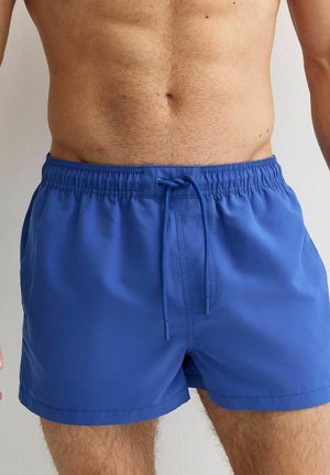 Męski tors ubrany w niebieskie kąpielówki sięgające do połowy uda, z elastycznym paskiem w talii i sznurkiem, stojący na jednolitym jasnym tle.