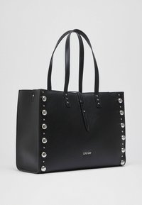 Sac cabas en cuir noir avec des clous décoratifs argentés, deux anses d'épaule et une fermeture éclair. Présente un logo embossé à l'avant.