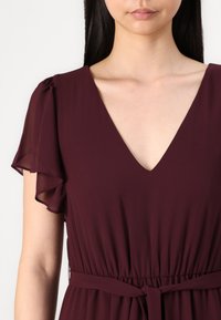 Burgundy dress in lightweight fabric with short, ruffled sleeves. Features a deep V-neck and a gathered waist with a tie detail.