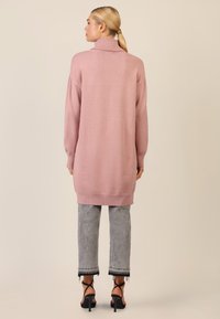 Langer rosa gestrickter Pullover mit hohem Kragen, gerippten Bündchen und Saum. Getragen über grauen cropped Jeans mit ausgefransten Enden und verzierten schwarzen Absatzschuhen.