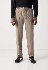 Pantalons beige à cordon de serrage dans une coupe décontractée, présentant une texture lisse et des plis subtils, associés à des chaussures de sport noires.