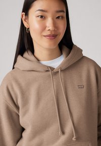 Hnedá mikina s kapucňou a šnúrkami, s prednou vreckom, vyrobená z mäkkého materiálu; má malé logo Levi's na hrudi.