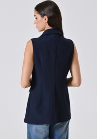 Marinefarbener ärmelloser Blazer mit taillierter Passform, versehen mit einem Reverskragen, glattem Stoff und einem leichten Schlitz am Rücken für Bewegungsfreiheit.
