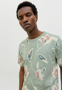 Šviesiai žalias marškinėliai su gėlėmis raštu, papuošti rožiniais, mėlynais ir kreminiais akcentais. Trumpi rankoviai ir apvalus iškirpimas. Lygi audinio tekstūra.