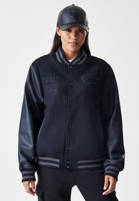 Mulher a usar uma jaqueta varsity preta dos Chicago Bulls com mangas de couro e um boné preto a condizer, de pé contra um fundo simples.