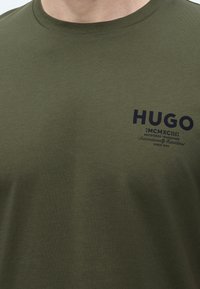 Λαδί πράσινο μπλουζάκι με μαύρο κείμενο "HUGO MCMXCIII Registered Trademark Internationally Established Since 1993" στο πάνω μέρος του στήθους.