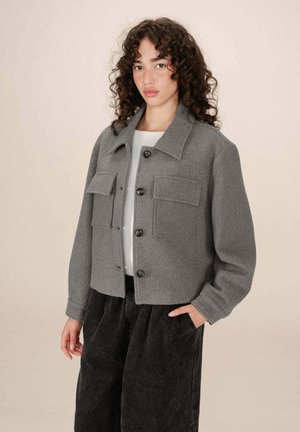 Veste courte en laine grise avec un col chemise, fermeture à boutons sur le devant et deux poches poitrine. Portée sur un haut blanc et un pantalon noir.