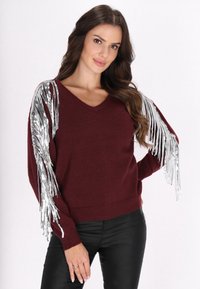 Pulover tricotat burgund cu decolteu în V și detalii cu franjuri argintii la mâneci, purtat cu pantaloni negri. Textură moale și croială lejeră.