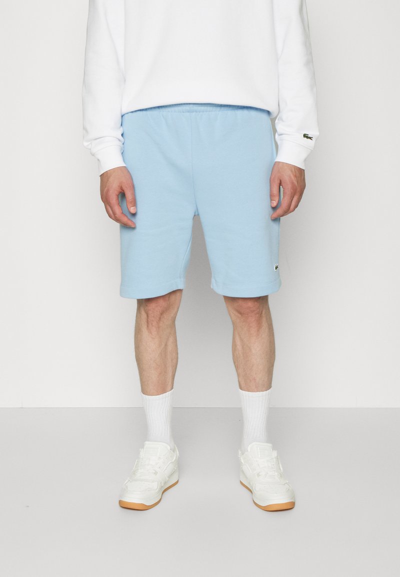 Shorts en coton bleu clair avec une taille élastique, poches latérales et détail de logo brodé. Associés à des chaussettes blanches et des baskets.