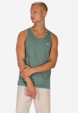 Uomo con capelli biondi corti che indossa una canottiera verde senza maniche e pantaloni beige, guarda a sinistra con le braccia incrociate davanti.
