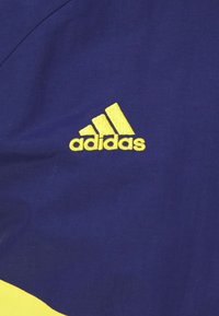 Logo Adidas v žluté barvě na tmavě modrém pozadí, s hladkým texturou a jednoduchým, moderním designem se třemi pruhy.