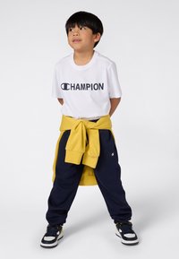 Hvit T-skjorte med "CHAMPION" logo i svart, marineblå joggebukser, gul hettegenser knyttet i livet, svarte og hvite sneakers.