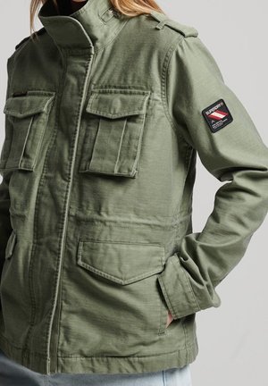 Grüne Utility-Jacke mit vier Klappentaschen, Reißverschluss und Knopfverschluss sowie einem Aufnäher am linken Ärmel, getragen von einer Person mit Hand in der Tasche.
