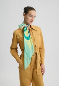 Tuta marrone chiaro con grandi tasche, una vita con cintura e una finitura texturizzata. Annodato attorno al collo c'è un foulard di seta a motivo teal e giallo.
