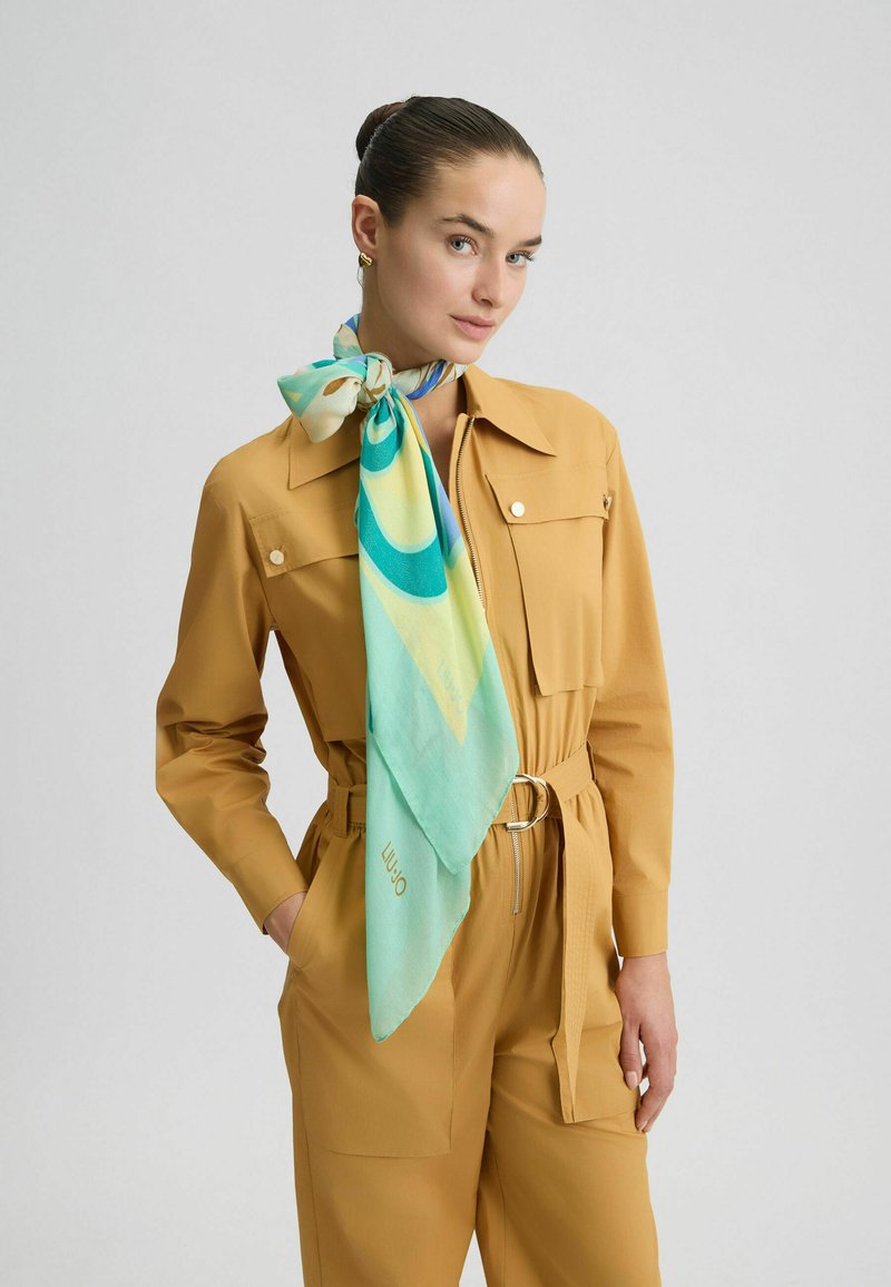 Lysbrun jumpsuit med store lommer, en bæltet talje og en struktureret finish. Bundet om halsen er et silkeskal med mønster i teal og gul.