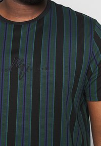 Camiseta negra con rayas verticales verdes y azules, usada por una persona, que presenta un sutil texto bordado en negro en el pecho.