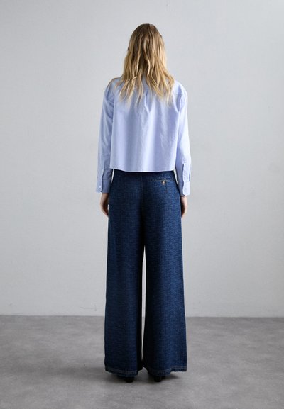 Γυναίκα με μακριά ξανθά μαλλιά, φορώντας ένα ανοιχτό μπλε κοντό μπλουζάκι και φαρδιά σκούρα μπλε παντελόνια με μοτίβο, ενώ στέκεται μπροστά σε έναν απλό γκρι τοίχο.