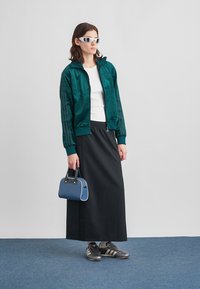 Green satin bomber jacket, white shirt, black skirt, blue handbag, and black athletic shoes with stripes. Model wears sunglasses.